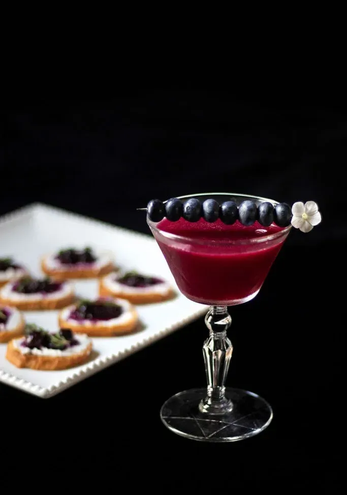 coquetel roxo escuro com mirtilos ao lado de um prato de aperitivos.' loading='lazy' title=