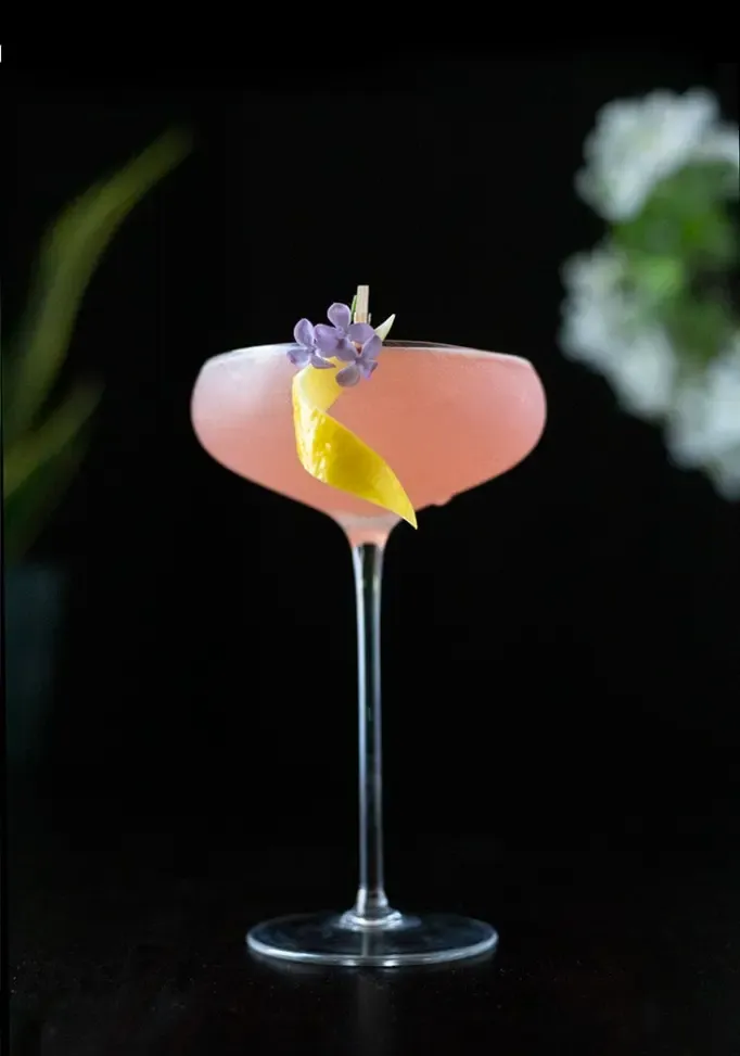 ein rosafarbener Cocktail auf schwarzem Hintergrund mit einer Zitronengarnitur und drei kleinen Fliederblüten' loading='lazy' title=