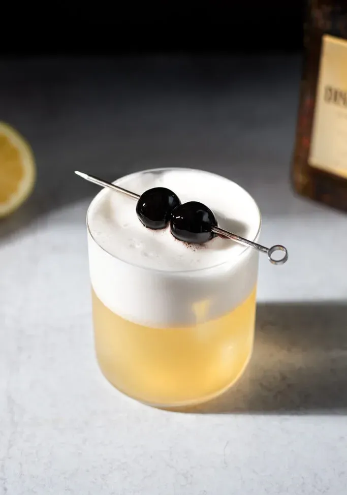 un cocktail doré avec une épaisse mousse blanche et deux cerises.' loading='eager' title=