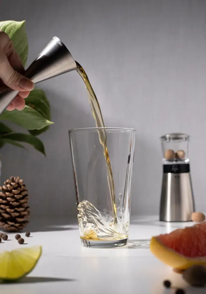 un jigger en métal versant du whisky dans un verre à mélange.' title=