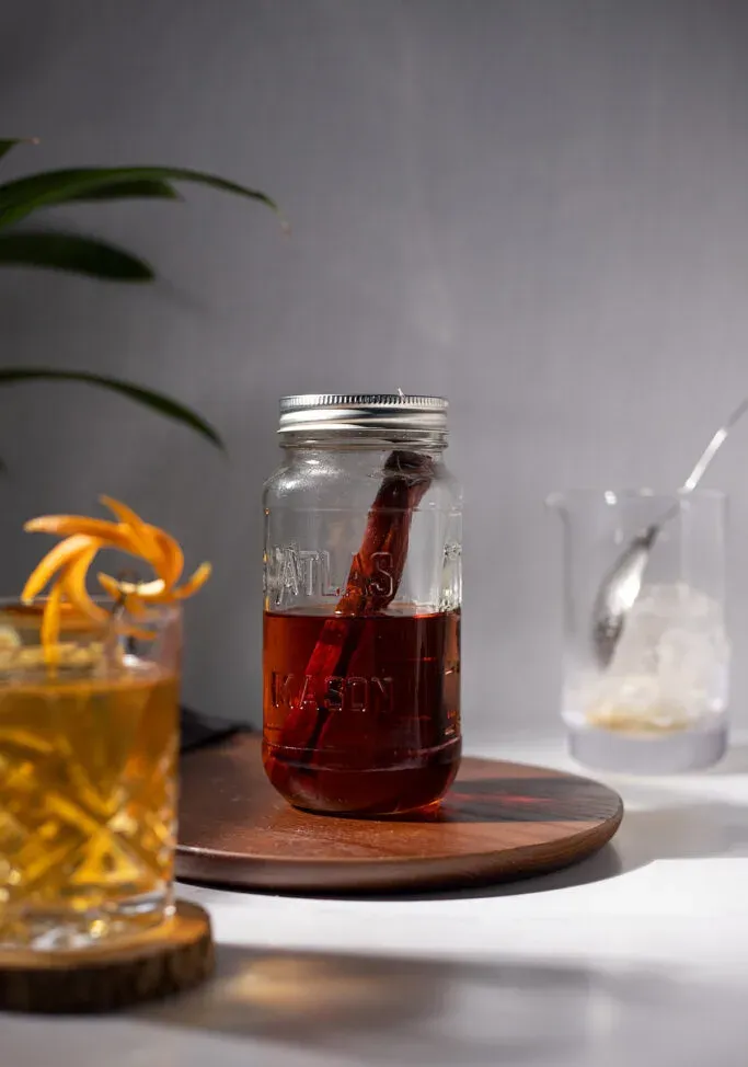 nádoba s tmavo hnedou alkoholom s paličkou z čerešňového dreva vedľa koktailu whisky a miešacie sklo.' title=