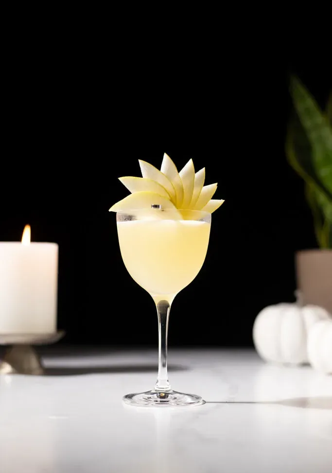 un cocktail jaune pâle dans un verre Nick et Nora avec une garniture en éventail de poire.' title=