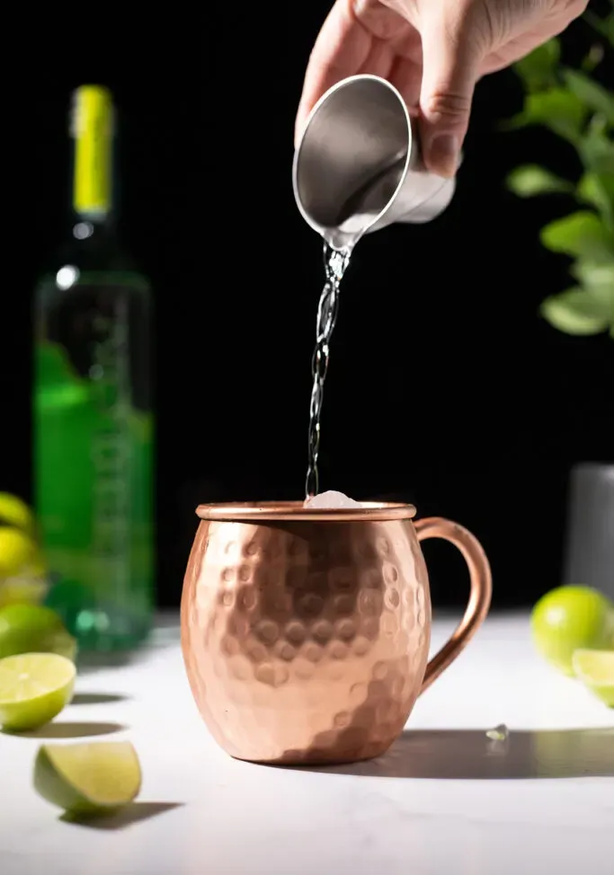 versando la cachaca trasparente da un jigger di metallo in una tazza di rame.' loading='lazy' title=