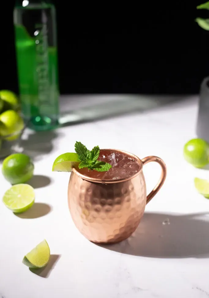 tazza di rame riempita con un cocktail brasiliano Buck accanto a lime e una bottiglia di Leblon Cachaca.' title=