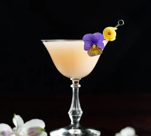 식용 꽃으로 장식된 옅은 크림색 칵테일입니다.' title=