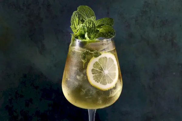 Close de um coquetel Hugo Spritz em uma taça de vinho, amarelo claro com uma rodela de limão na lateral da taça e um grande buquê de hortelã