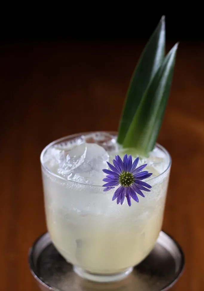 une boisson jaune pâle garnie d'une fleur violette et de feuilles d'ananas.' loading='lazy' title=