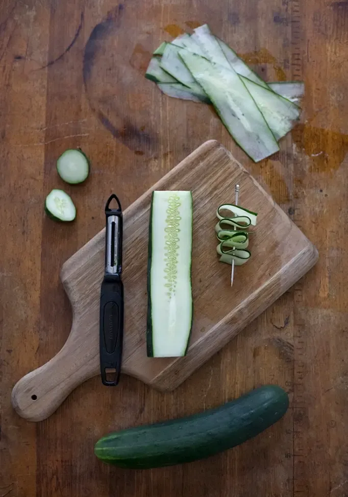 pepinos, um descascador de legumes e um palito de coquetel com fitas de pepino espetadas nele.' title=