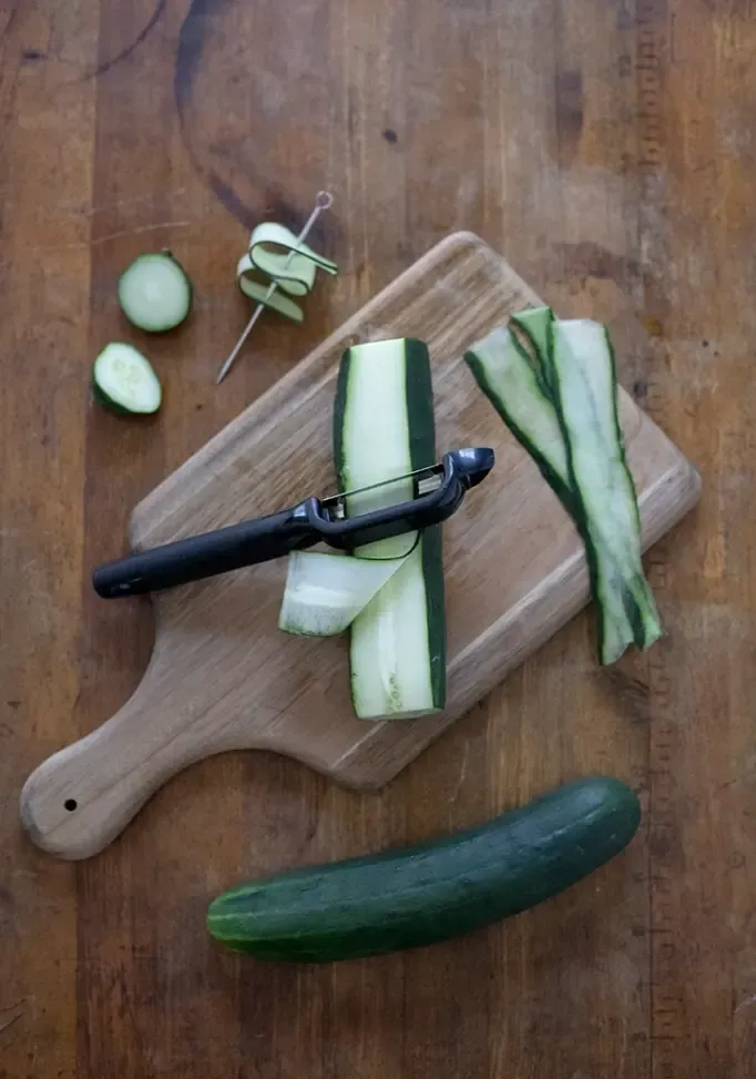 descascar uma camada de pepino com um descascador de legumes para fazer uma guarnição de fita de pepino' title=