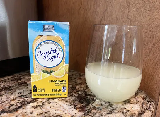 A container of Crystal Light brand lemonade packets next to a small glass of the beverage.