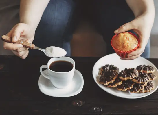 woman pouring sugar into coffee, holding muffin above plate of cookies