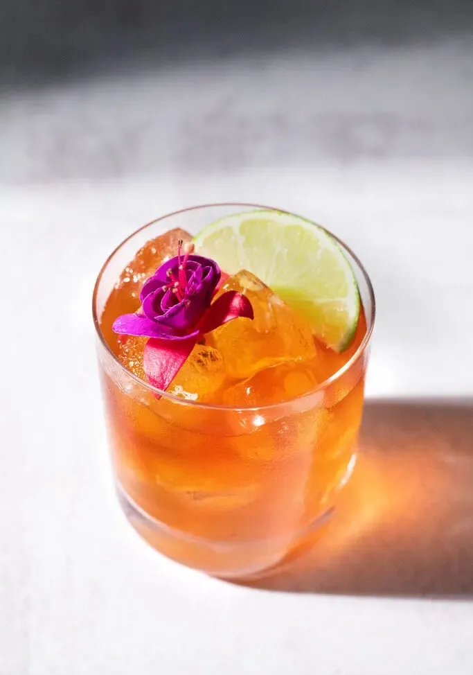 un cocktail marrone in un bicchiere rock con un fiore e uno spicchio di lime.' title=