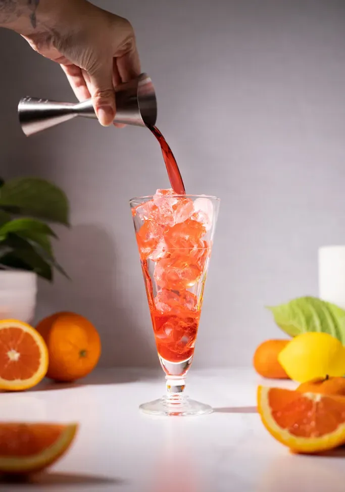 nalewanie Campari ze srebrnego jiggera do wysokiej szklanki z lodem.' loading='lazy' title=