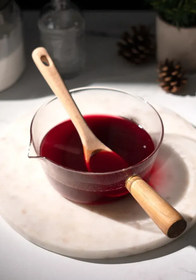 cucchiaio di legno appoggiato in un pentolino di vetro pieno di liquido rosso scuro.' title=