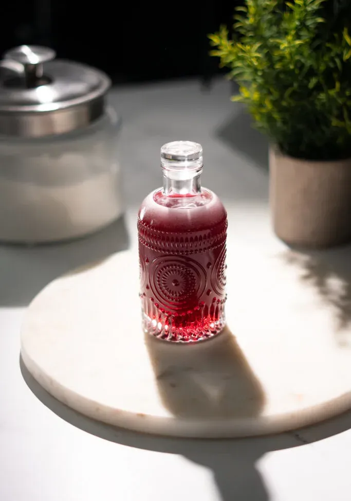 piccola bottiglia di vetro di liquido rosso su un tavolo con una pianta e un barattolo di zucchero.' title=