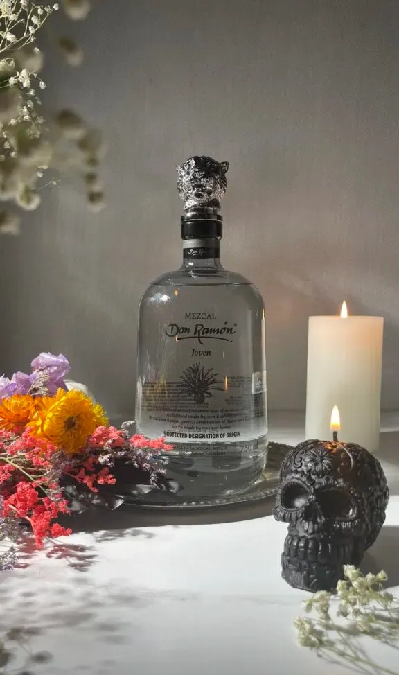 láhev Don Ramon mezcal vedle svíček a květin.' loading='lazy' title=