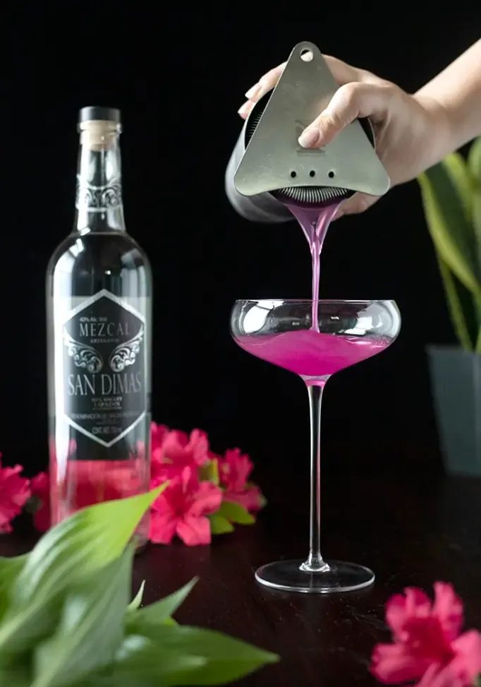 versando un cocktail rosa dallo shaker in un bicchiere alto da coupé' loading='lazy' title=