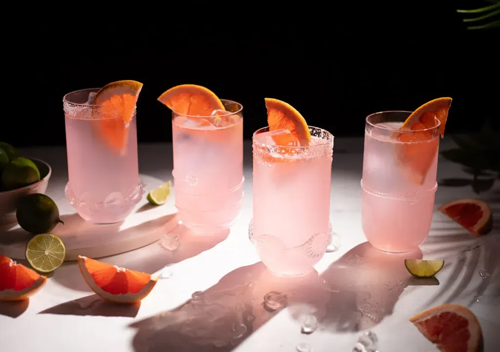 vier mit rosa Flüssigkeit gefüllte Highball-Gläser und Grapefruitspalten.' loading='lazy' title=