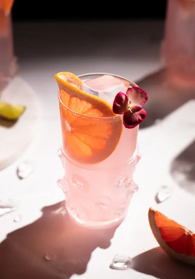 Rosa Getränk mit klarem Eis, Grapefruitrad und kleiner rosa Blume.' loading='lazy' title=