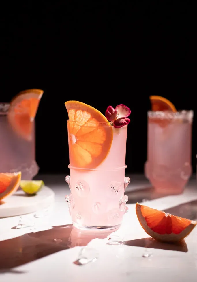Drei rosa Getränke mit Grapefruitgarnitur auf weißem Tisch in der Sonne.' title=