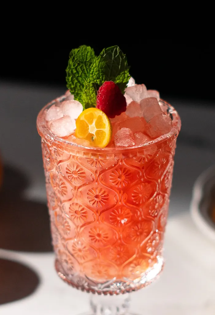 Cerca de líquido rojo en un vaso grabado con frambuesa y hojas de menta.' title=