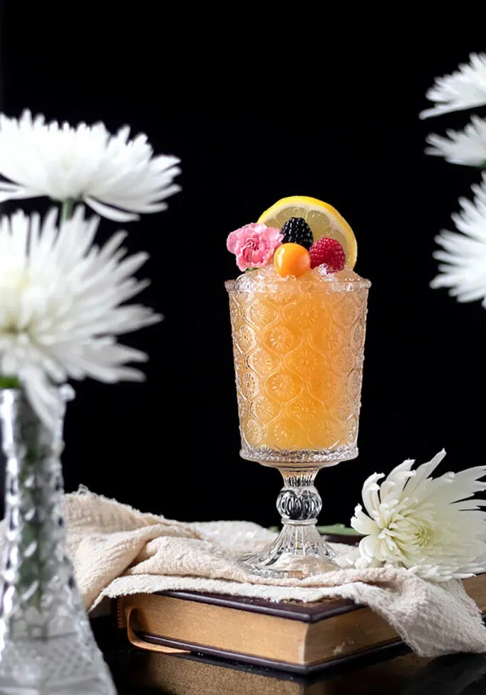 Bebida de color naranja pálido en una copa de cristal sobre un libro junto a flores.' title=