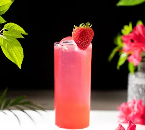 ein leuchtend rosa Getränk in einem hohen Glas neben rosa Blumen.' title=