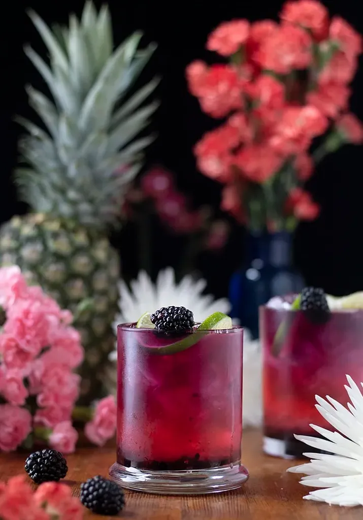 パイナップルと明るいピンクの花の隣にブラックベリーをトッピングした 2 つの濃い赤色のドリンク。' loading='lazy' title=