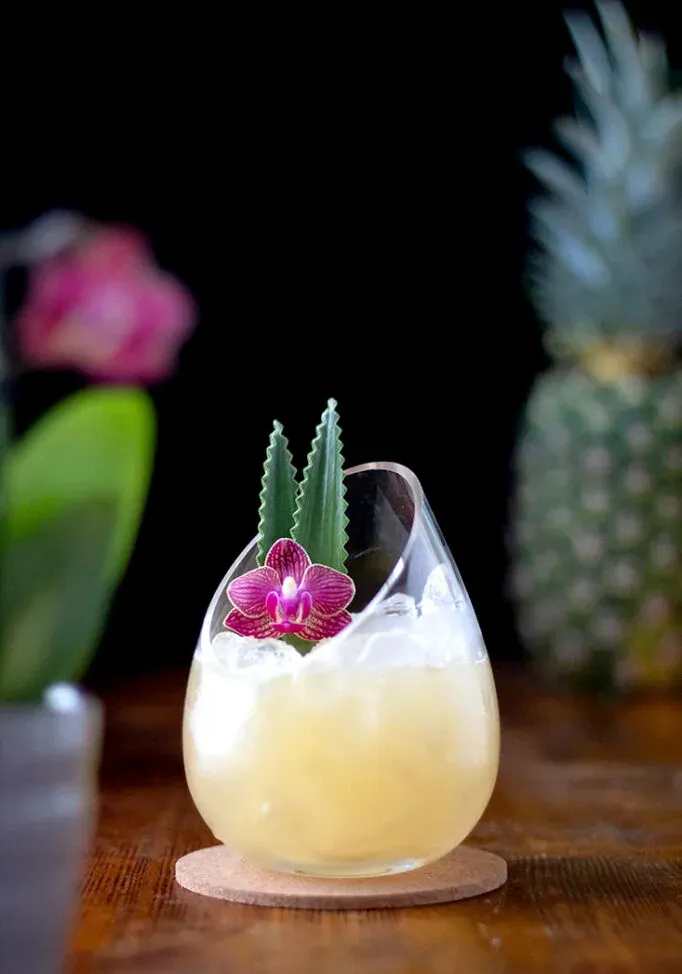 bevanda giallo pallido guarnita con un'orchidea rosa accanto a un ananas.' loading='lazy' title=