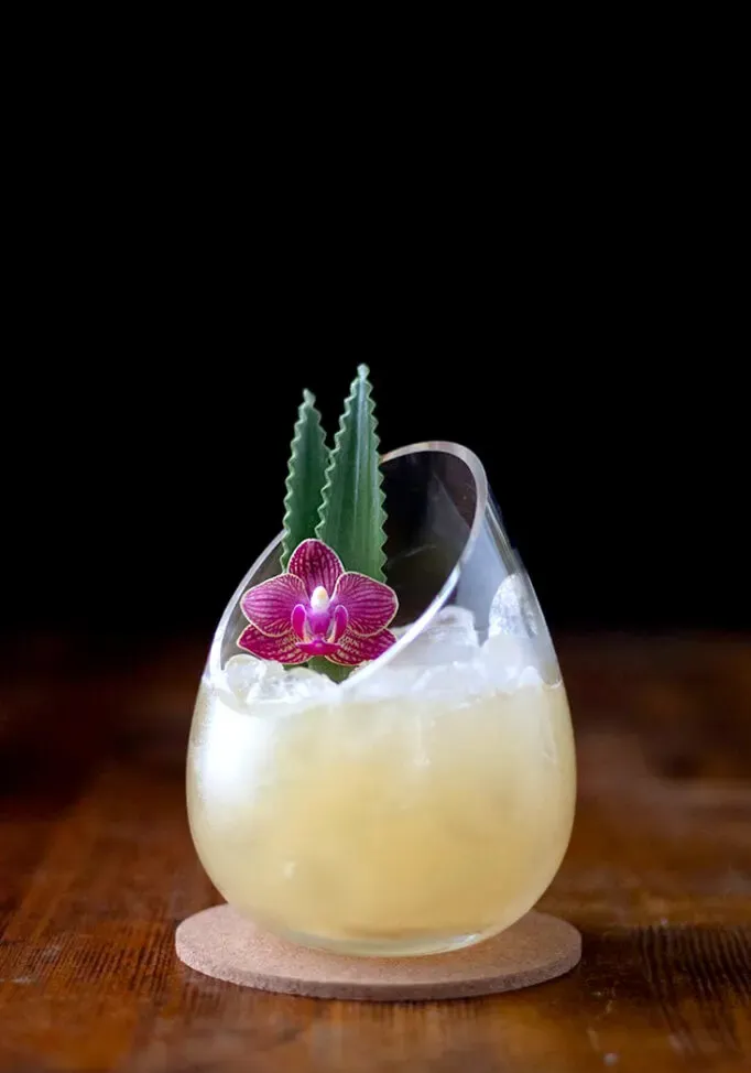 bevanda giallo pallido in un elegante bicchiere moderno con foglie di ananas e un'orchidea.' loading='lazy' title=