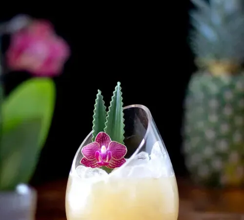 bevanda giallo pallido guarnita con un'orchidea rosa accanto a un ananas.' title=