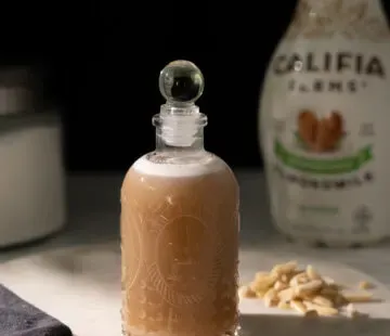 malá skleněná láhev světle hnědého sirupu vedle bílé láhve mandlového mléka Califia.' title=