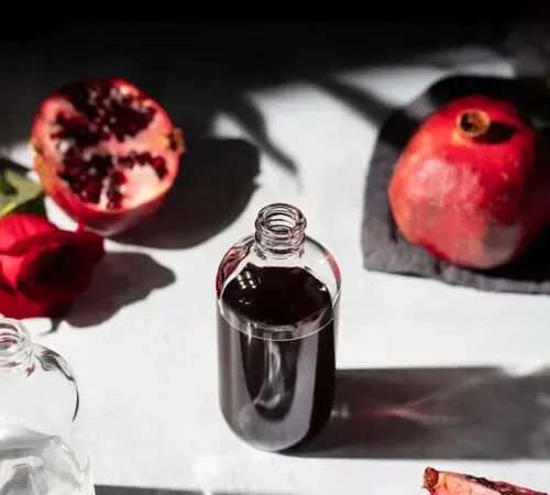 eine kleine Apothekerflasche mit dunkelroter Flüssigkeit, umgeben von Granatäpfeln' title=