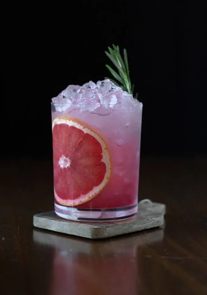ein rosa Getränk in einem Steinglas mit einem roten Grapefruitrad und einem Rosmarinzweig.' title=