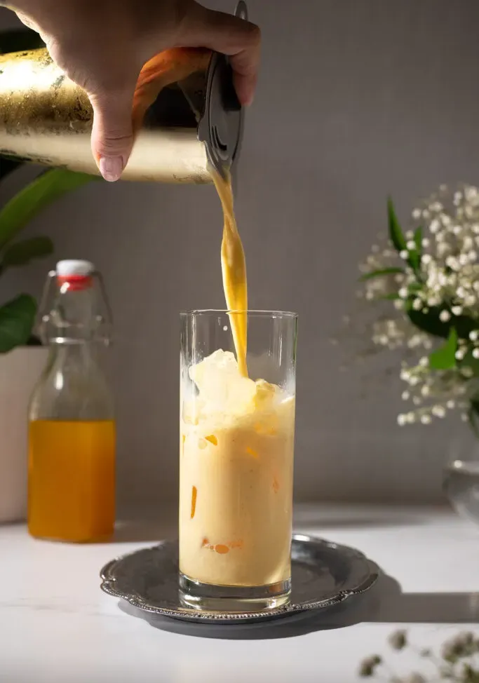 versando il latte dorato da uno shaker in un bicchiere pieno di ghiaccio.' loading='lazy' title=