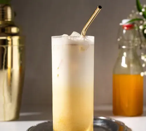 un bicchiere di latte dorato ghiacciato accanto a uno shaker dorato e una bottiglia di sciroppo di curcuma.' title=
