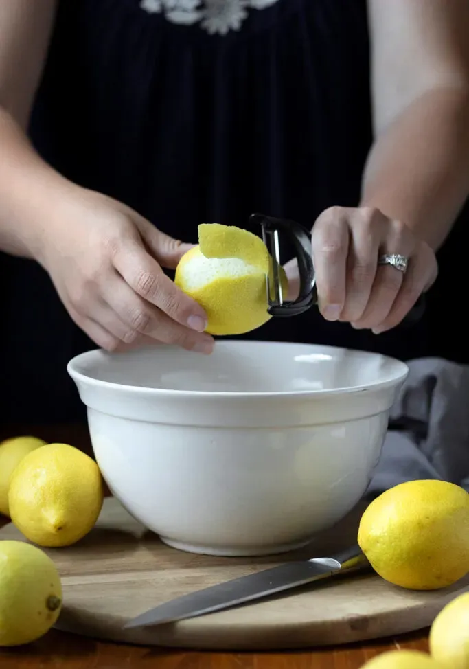 utilizzando un pelapatate per rimuovere le bucce di limone sopra una ciotola.' loading='lazy' title=