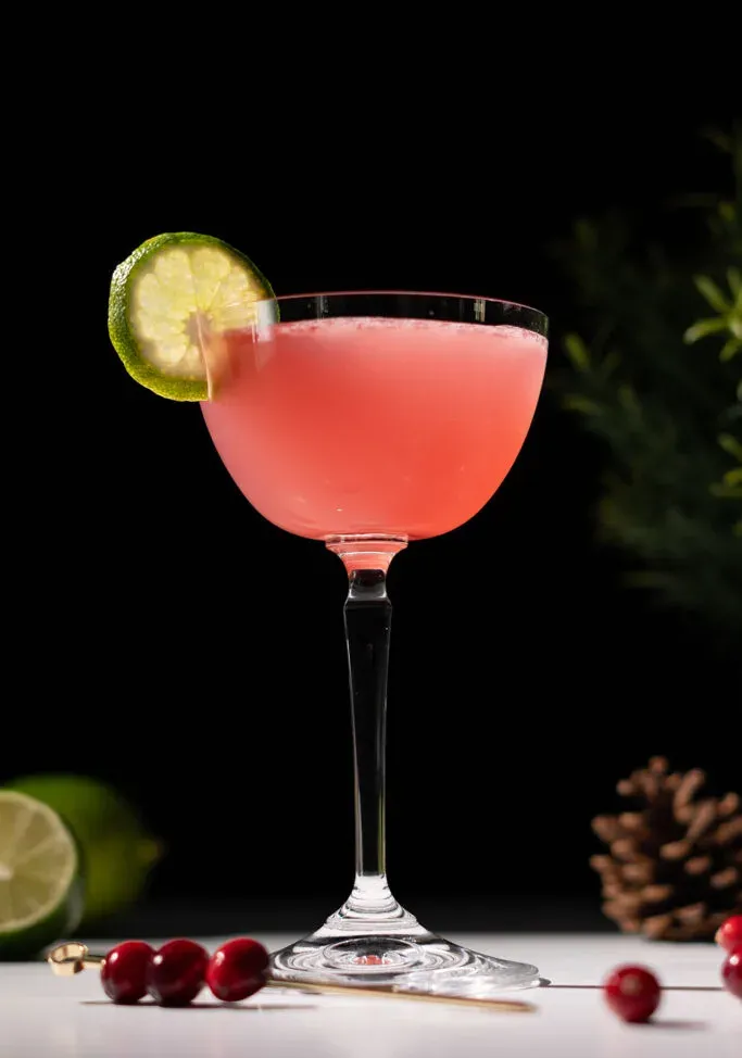 primo piano di un cocktail rosso in un bicchiere da coupé con una ruota di lime sul bordo.' title=