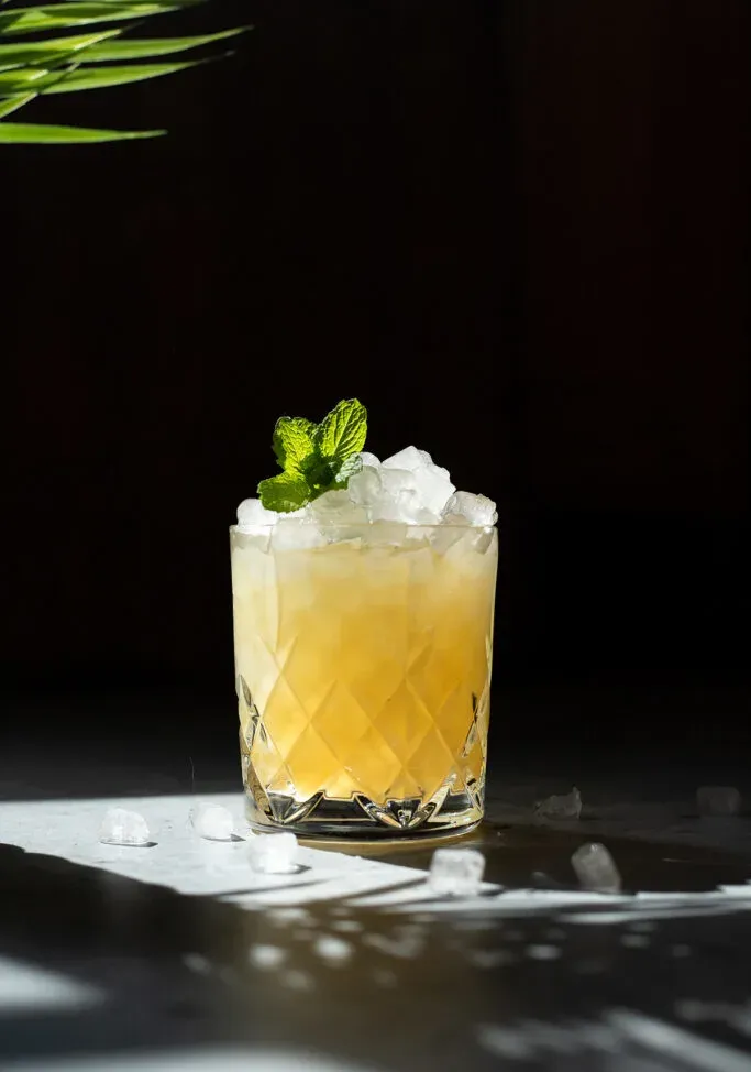 un cóctel amarillo con hielo picado en un vaso bajo adornado con menta.' title=
