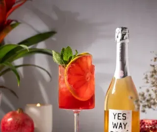 kieliszek do wina wypełniony jasnoczerwonym napojem i koło grejpfrutowe.' title=