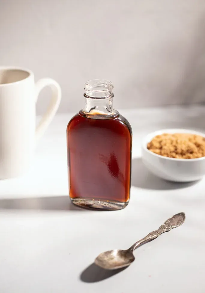 une petite bouteille en verre de sirop brun foncé à côté d'une tasse de café blanche.' loading='lazy' title=
