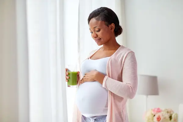 pregnant african american woman drinking green vegetable juice or smoothie at home