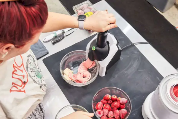 Osoba, ktorá tlačí na vrch servírovača Yonanas Frozen Fruit Soft Serve Maker, zatiaľ čo ružové mäkké servírovanie dávkuje do sklenenej misy
