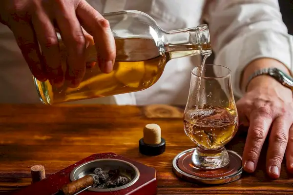 Eine Person gießt Whisky in ein Glas auf einem Holztisch neben einer Zigarre