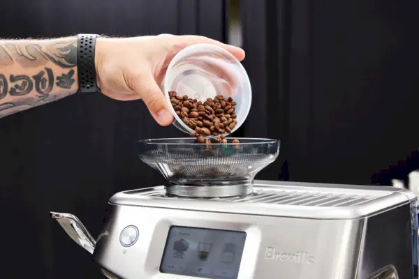 Uma pessoa despejando grãos de café no moedor de café conectado ao Breville The Barista Touch Impress (BES881BSS1BNA1)