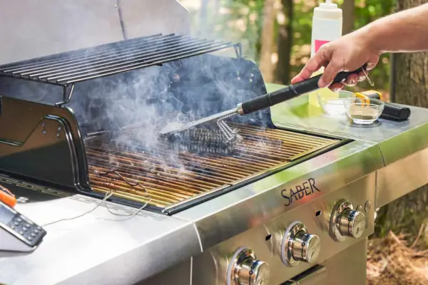 Uma pessoa usa a escova para grelhar para limpar a churrasqueira a gás propano líquido de aço inoxidável com 3 queimadores Sabre Select