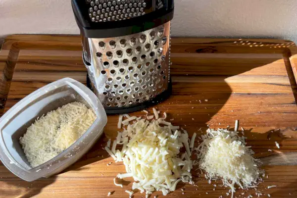 Nerezové škatuľové strúhadlo KitchenAid Gourmet s odnímateľnou odkladacou nádobou umiestnenou na drevenom stole s plátkovým syrom
