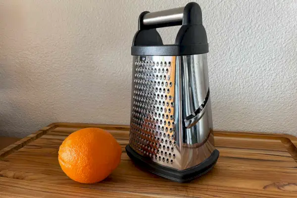 Nerezové škatuľové strúhadlo KitchenAid Gourmet s odnímateľnou úložnou nádobou je vystavené na drevenom stole vedľa pomaranča