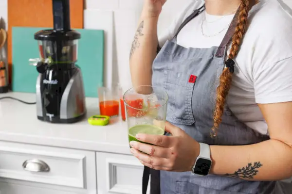 Osoba, ktorá drží pohár zeleného džúsu pred odšťavovačom Tribest Slowstar Vertical Cold Press Juicer
