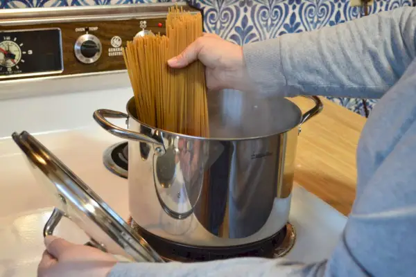 Eine Person gibt trockene Spaghetti in einen Mueller UltraClad Tri-Ply Edelstahl-Suppentopf mit Deckel und Schöpflöffel, 8 Quart